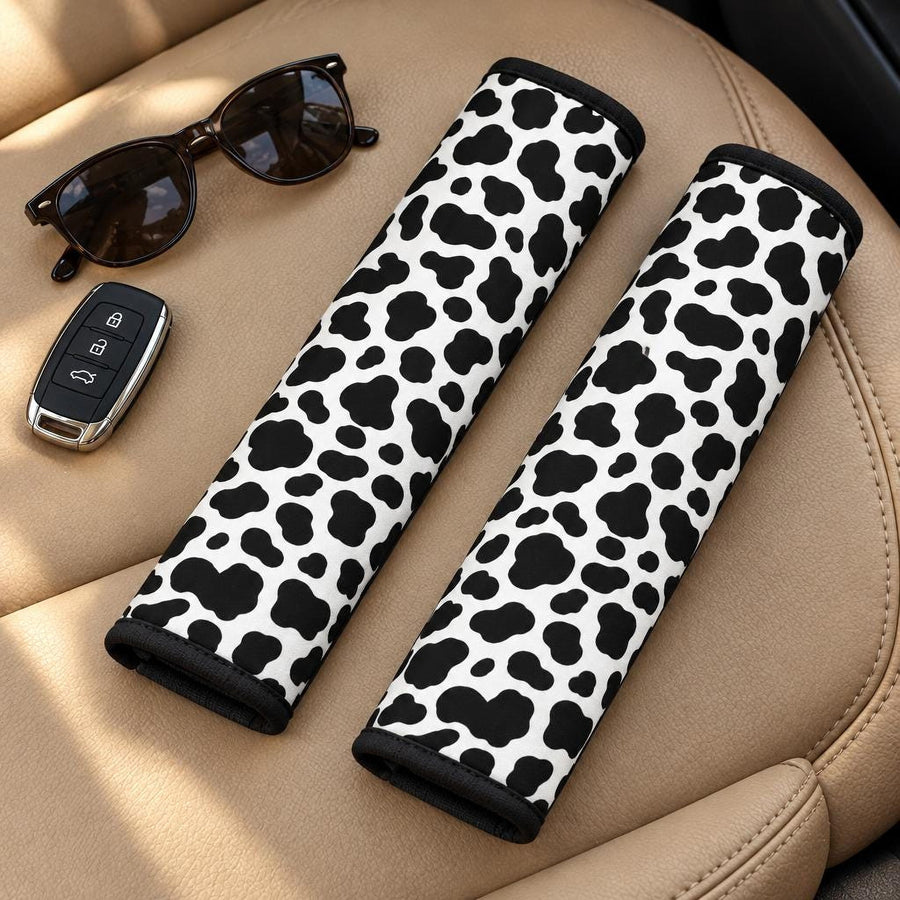 Cow Print Seat Belt Covers (Set of 2) lying side by side on a tan leather car seat with sunglasses and a car key